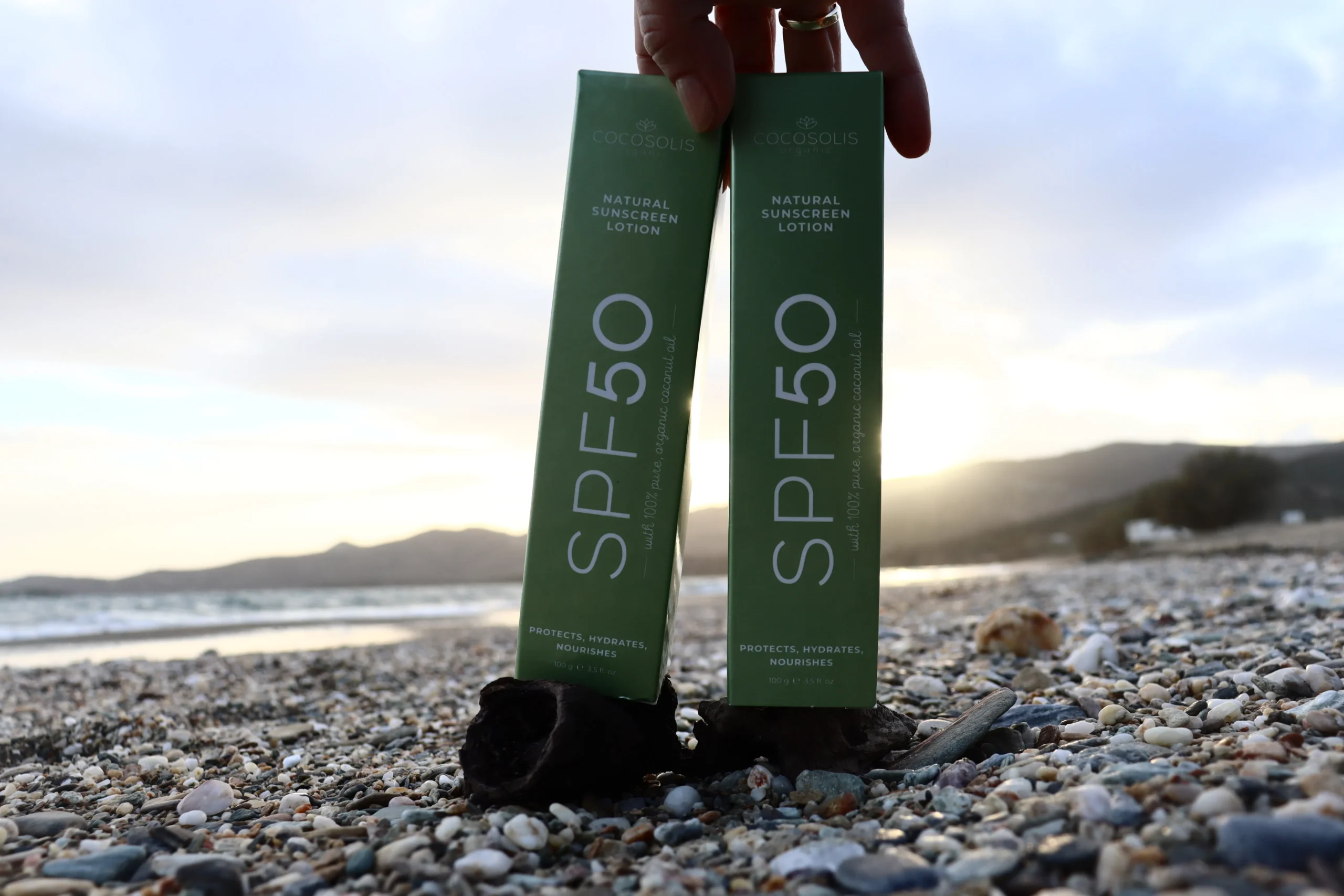 Two SPF50 sunscreen lotion tubes on a rocky beach at sunset, promoting smooth skin benefits.