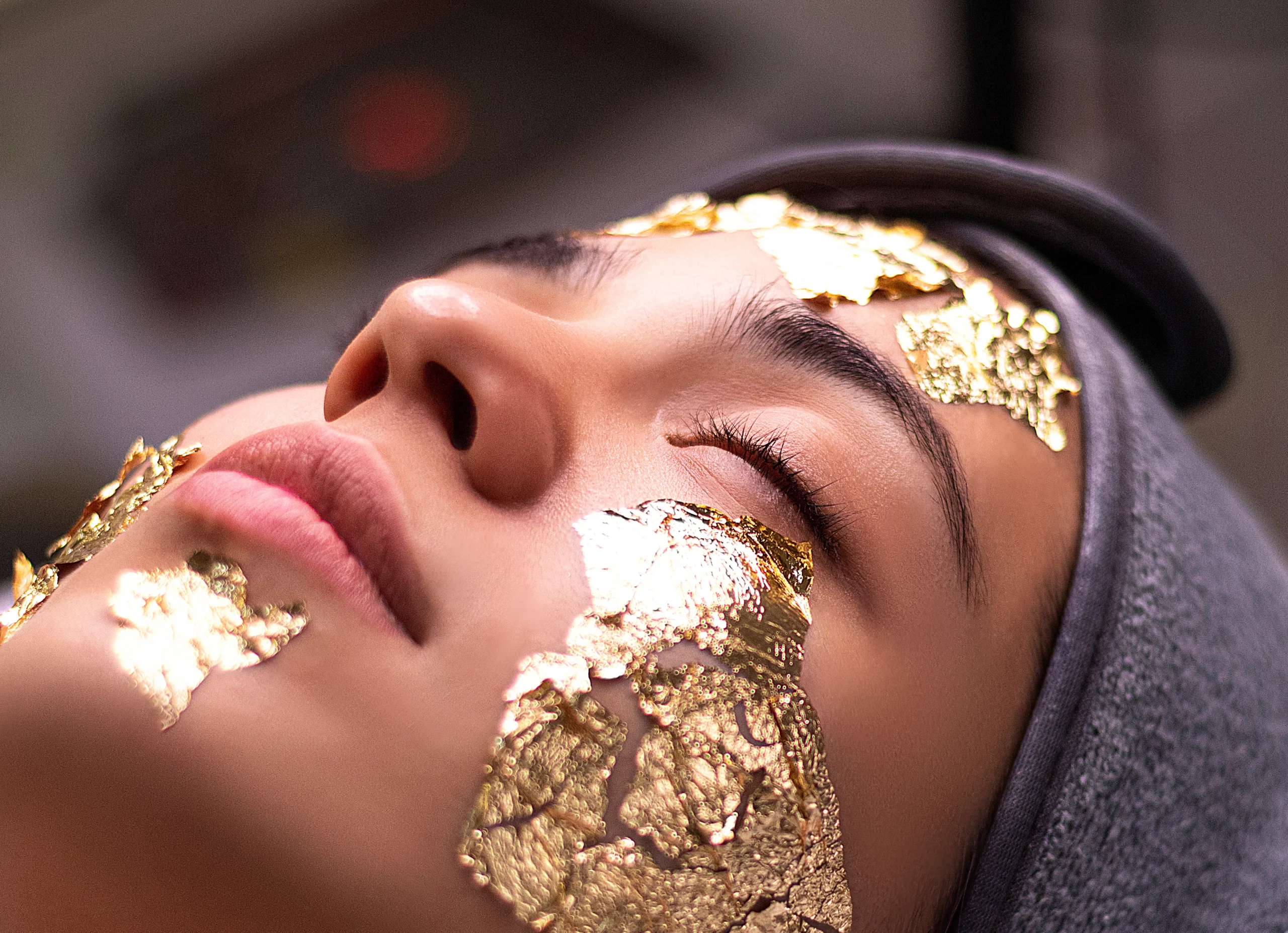 Close-up of a woman's face covered in gold leaf during a beauty treatment, possibly a luxurious skincare procedure.