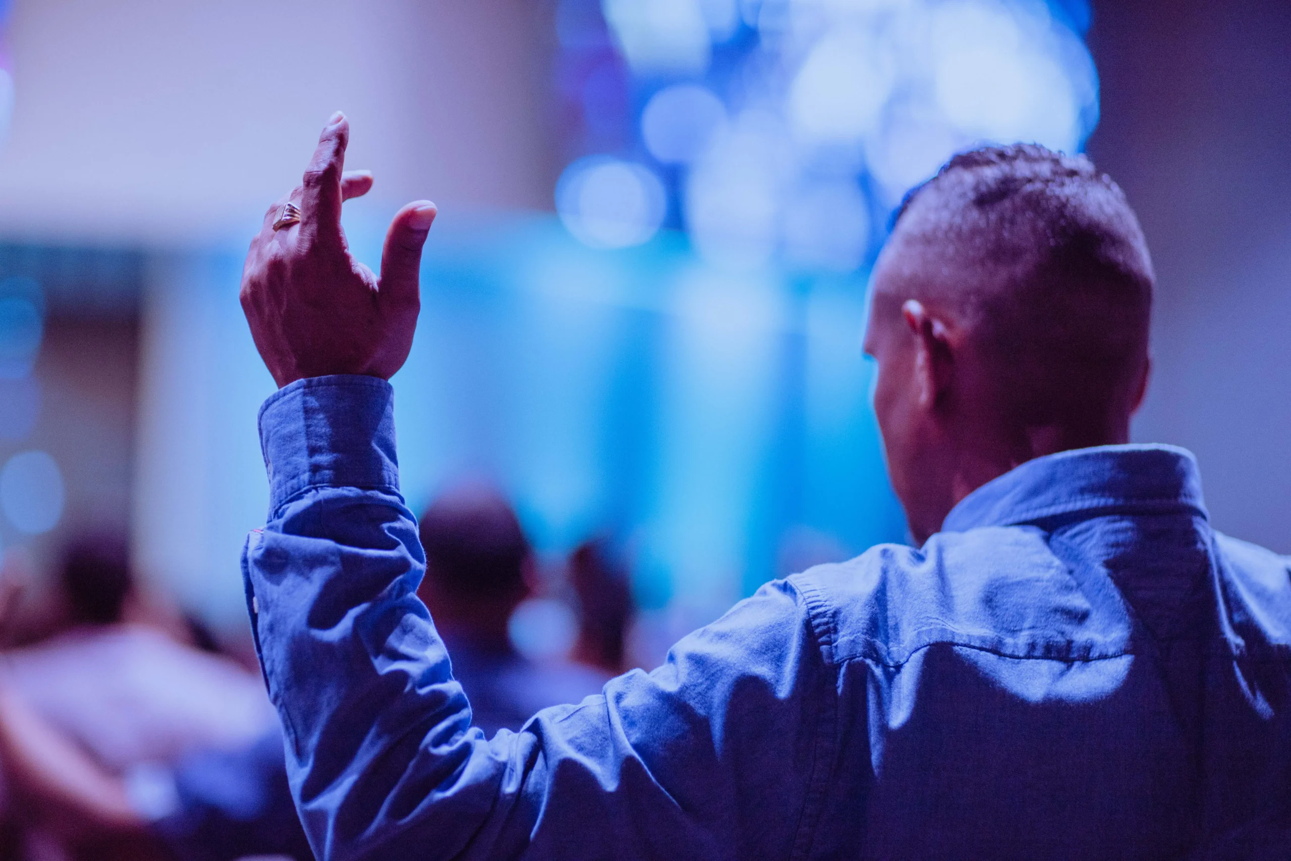 Man in blue shirt with raised hand in a crowd with blue lighting.