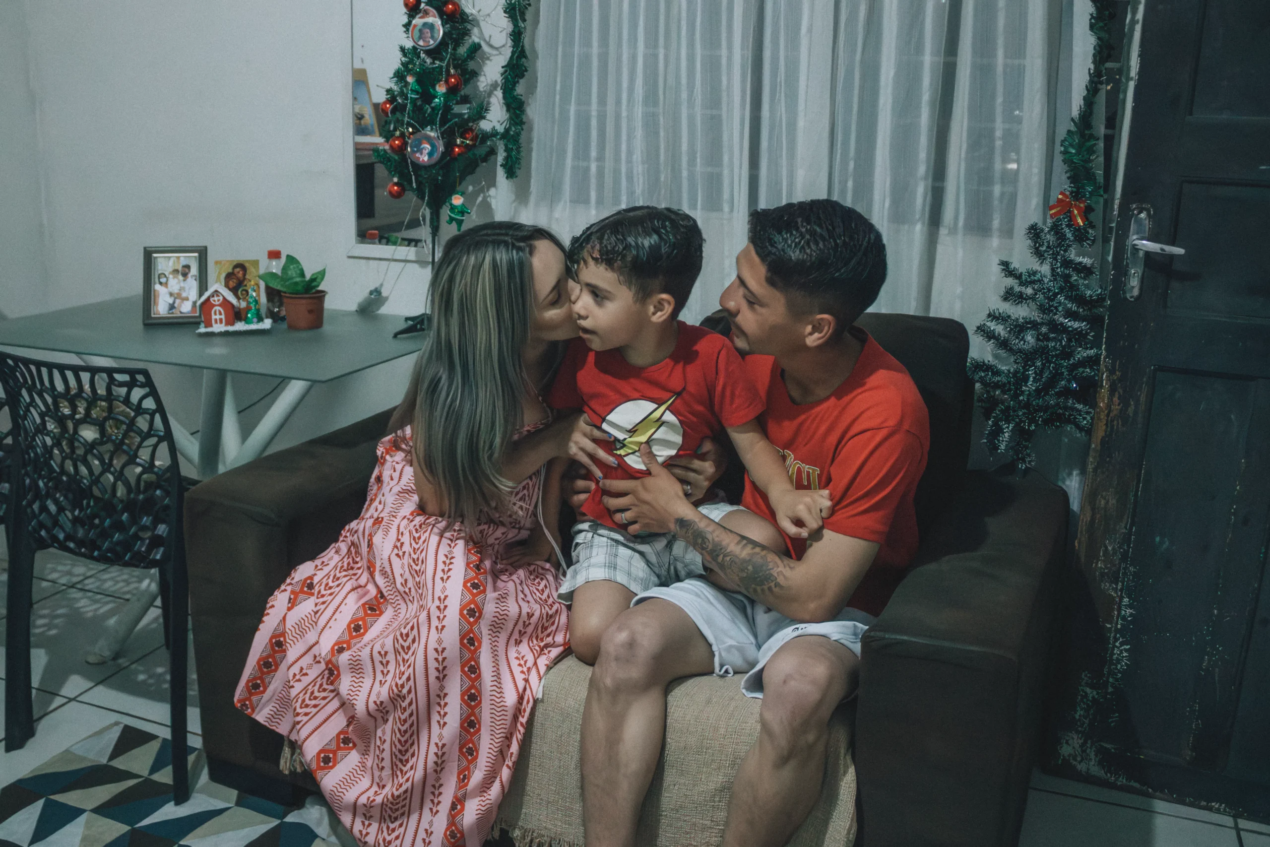 Family of three in red shirts sitting on couch near Christmas decor.
