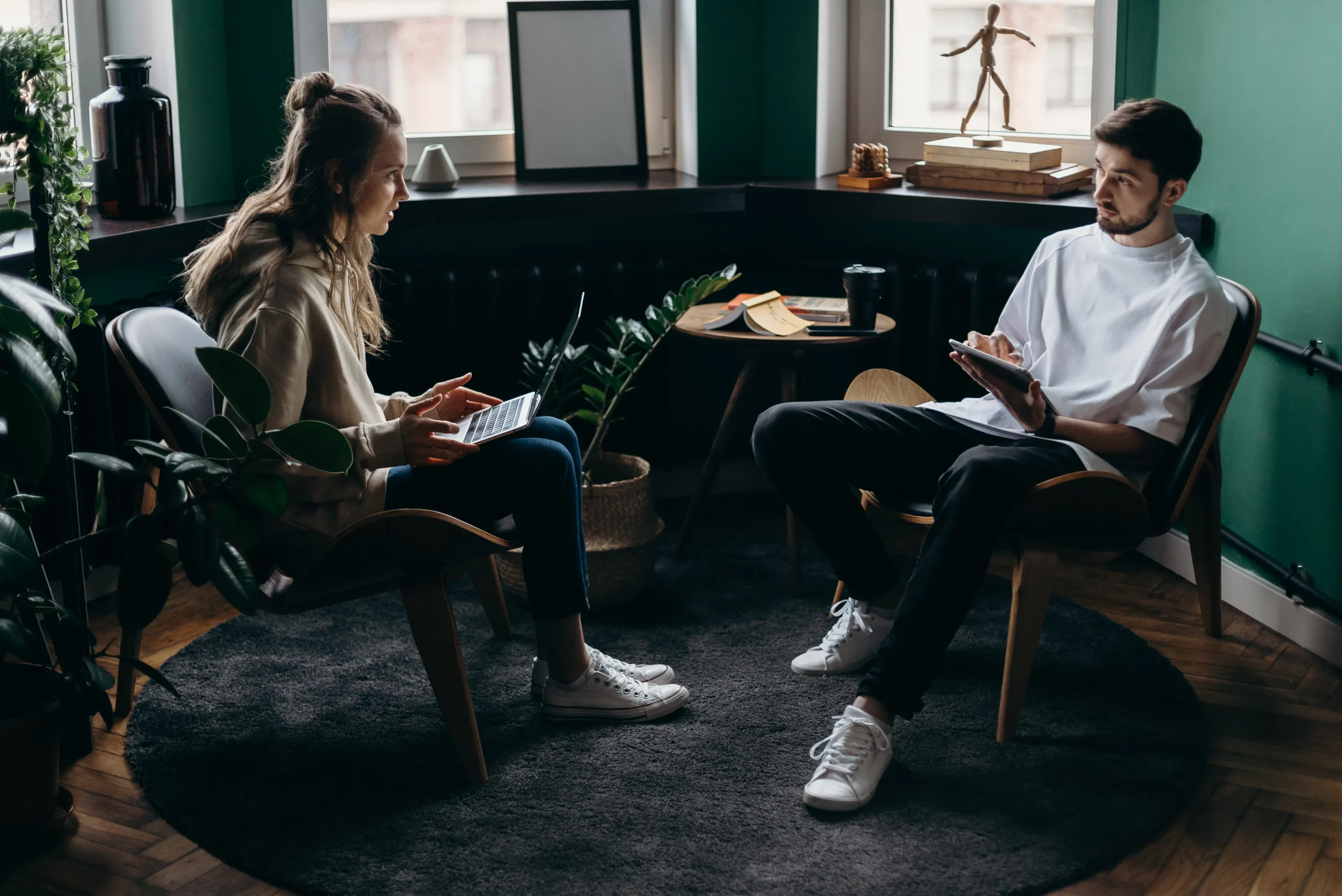 Two people in discussion, one using a laptop, in a bright room with plants.