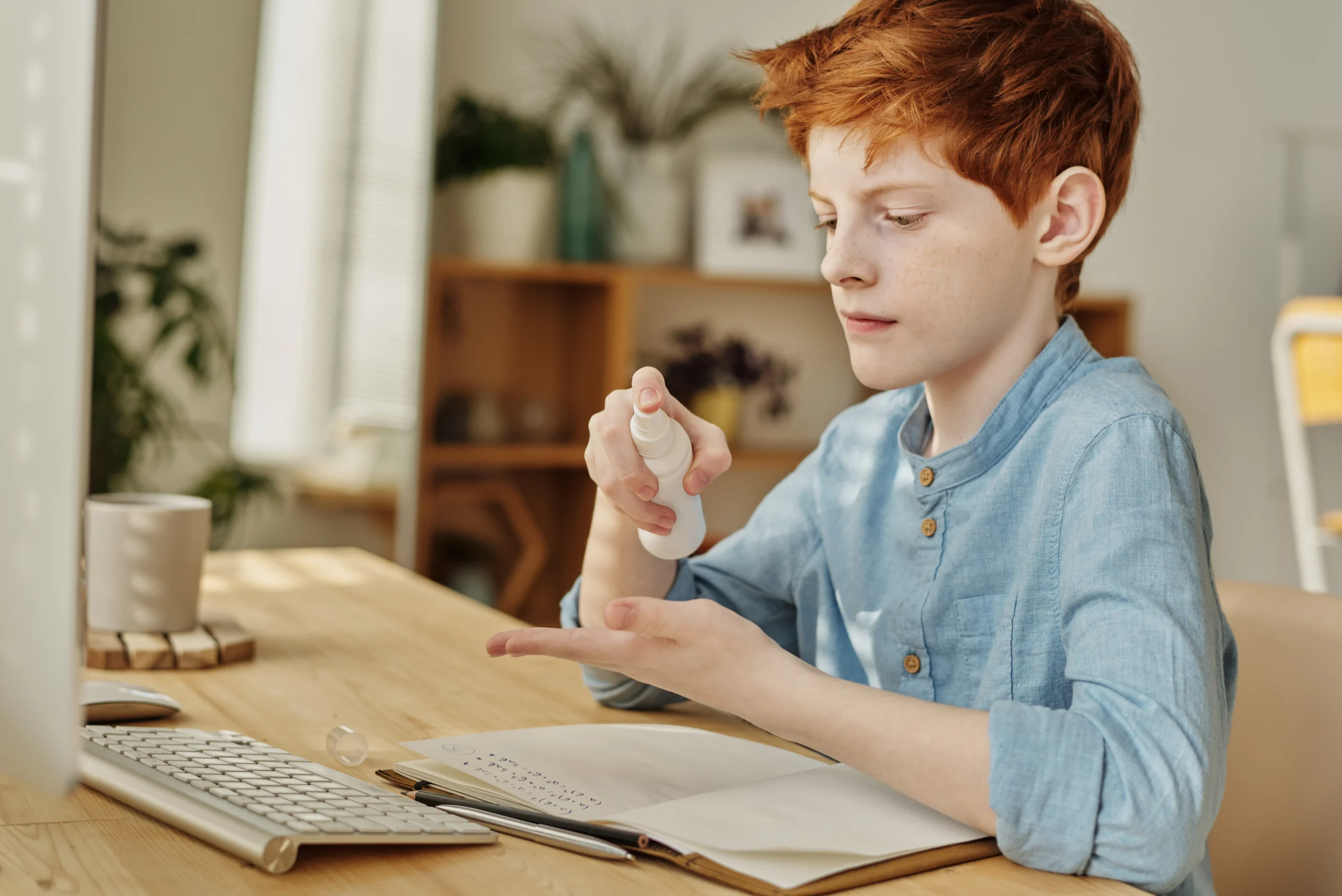 Red-haired boy sanitizing hands at desk with computer.