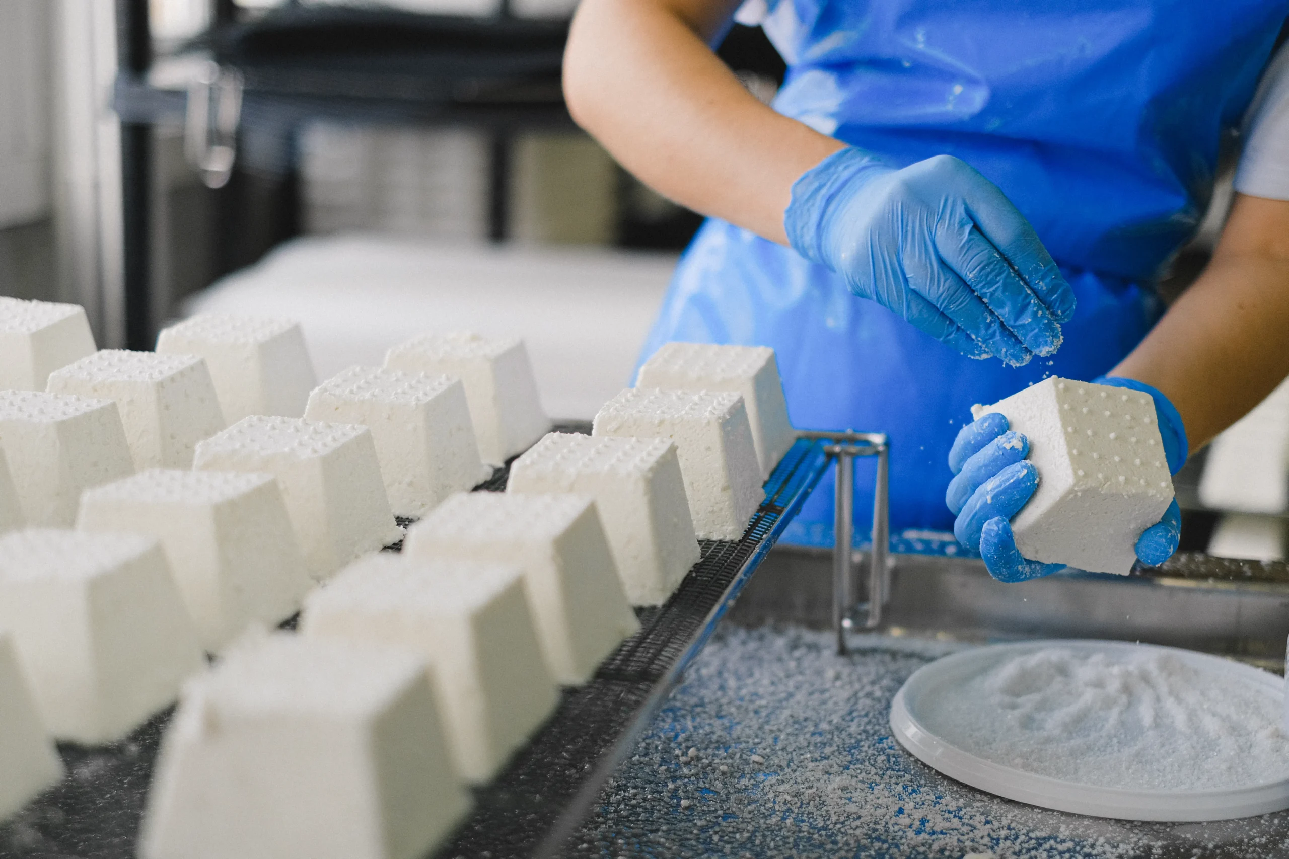 Gloved worker handling blocks of white cheese in a food processing plant.