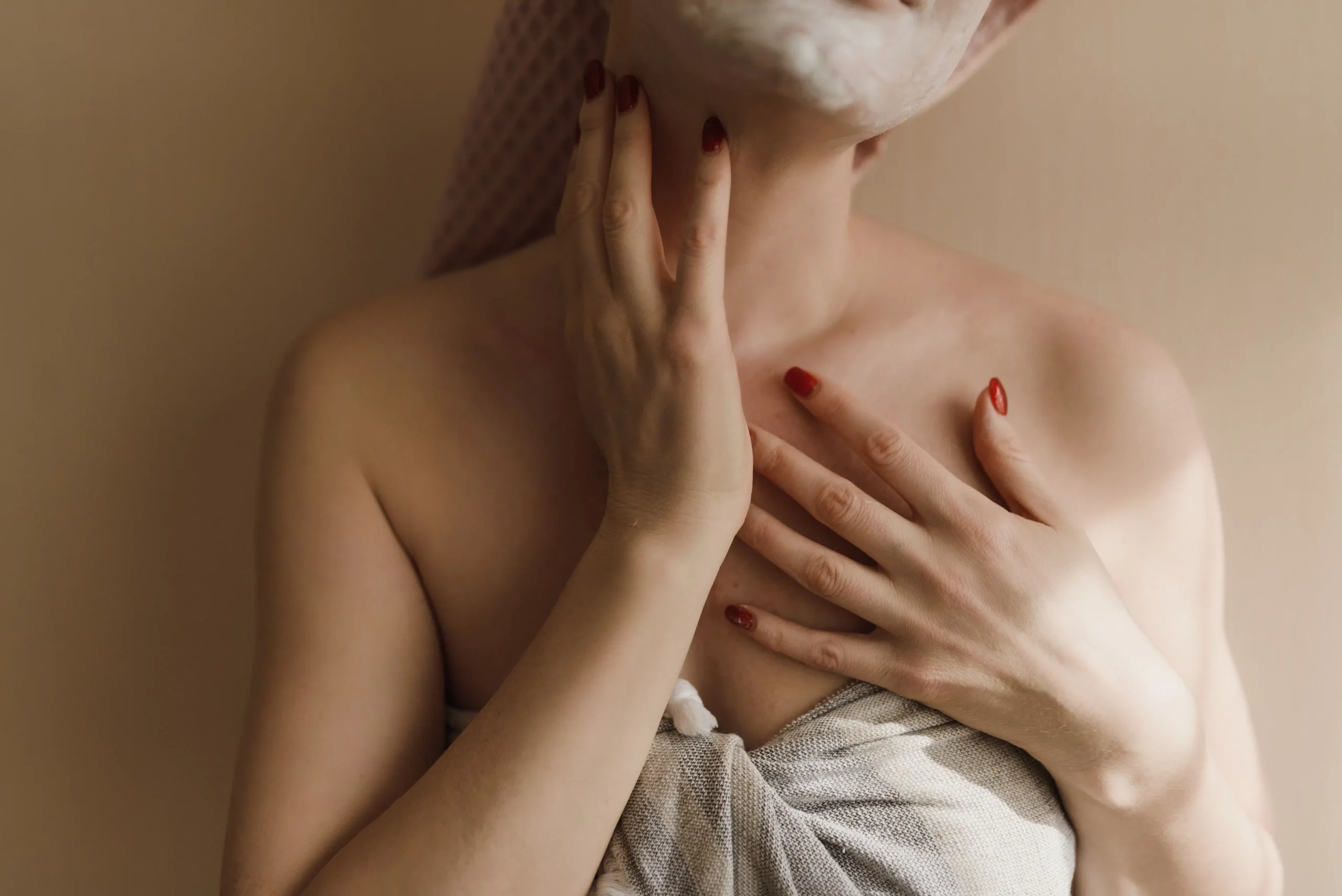 A woman with a face mask and towel on her head, gently touching her neck and chest.