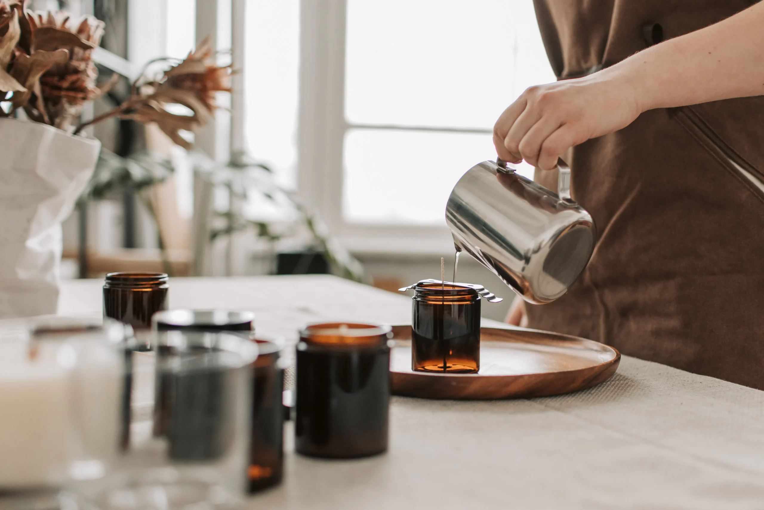 A person in an apron carefully pours hot wax into amber glass jars, possibly for candle making.
