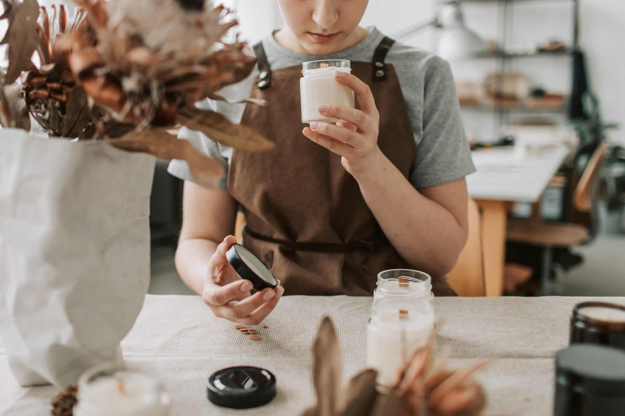 Person inspecting a candle in a jar, with crafting materials on a table.