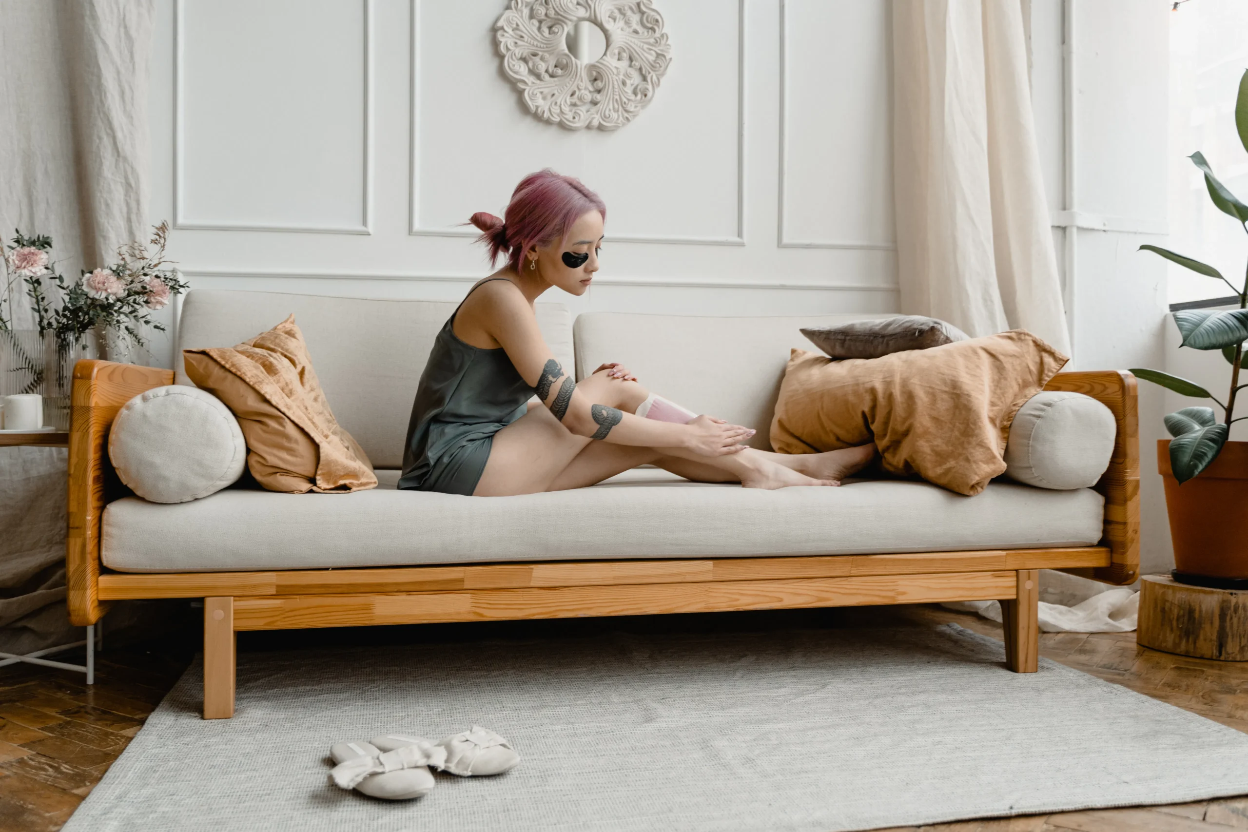 Woman with pink hair applying leg wax on a couch for smooth legs.