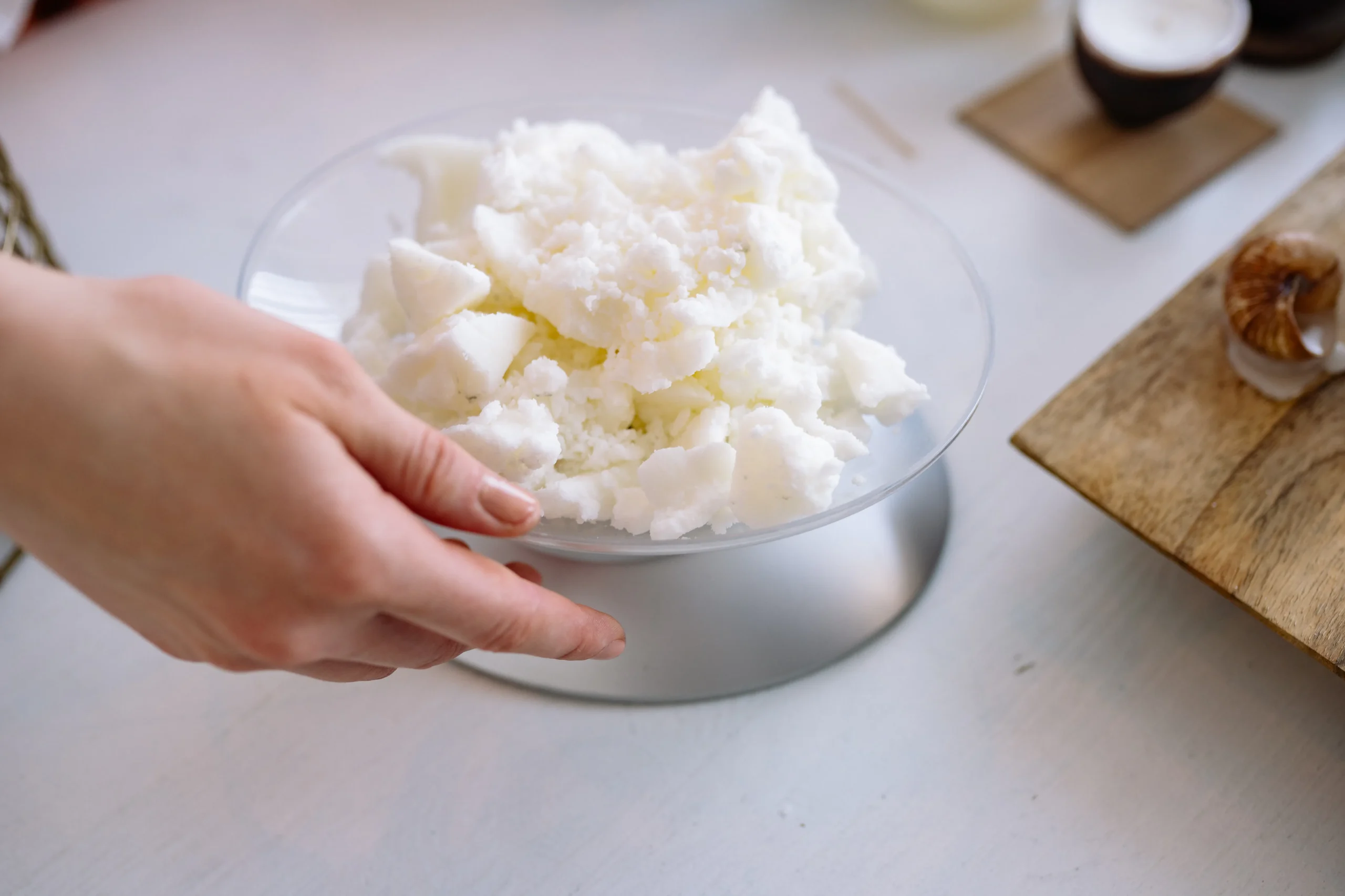 A close-up of a hand holding a clear bowl filled with white shea butter, likely for use in skincare or cosmetic products.