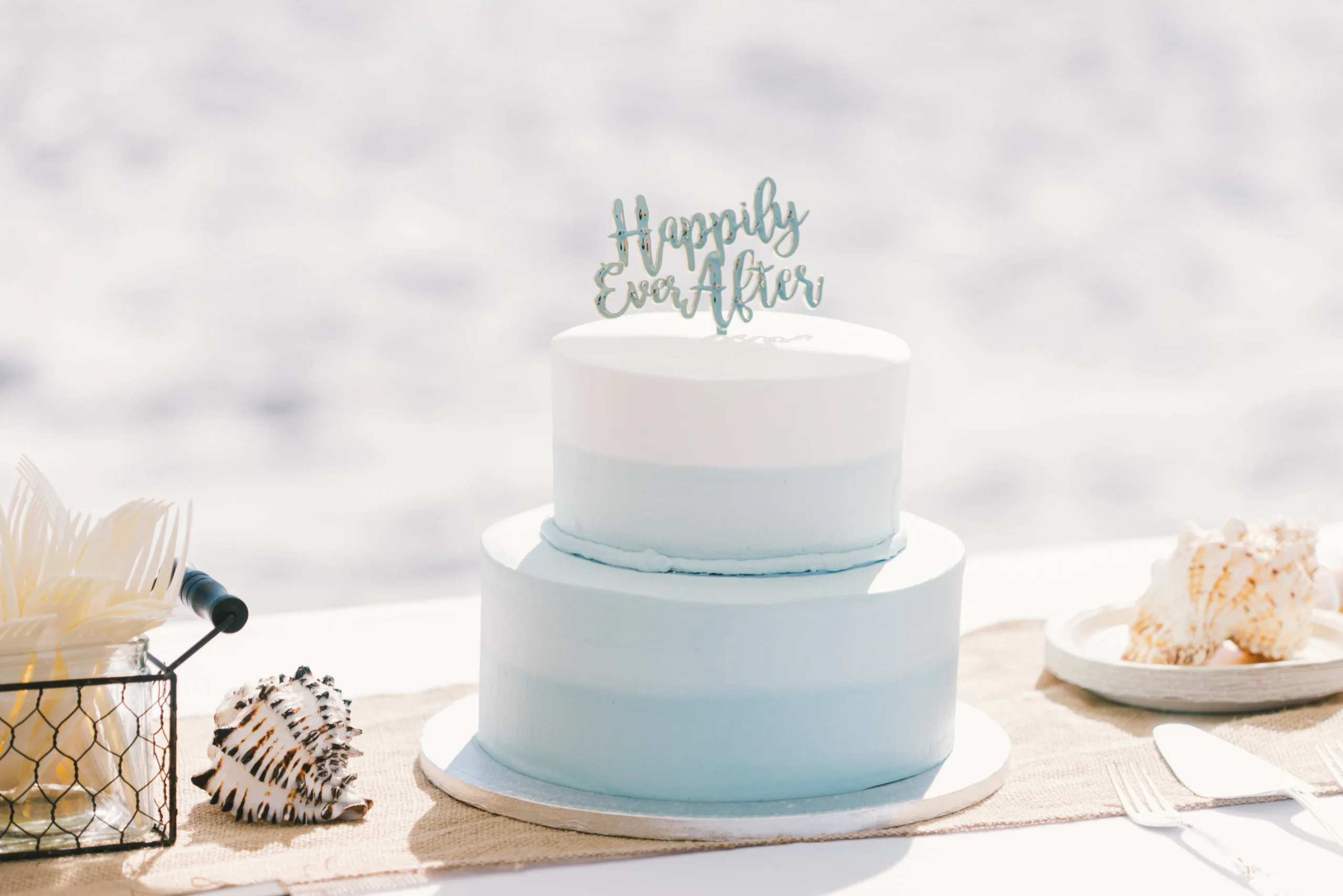 Two-tiered cake with 'Happily Ever After' topper, beach wedding theme.