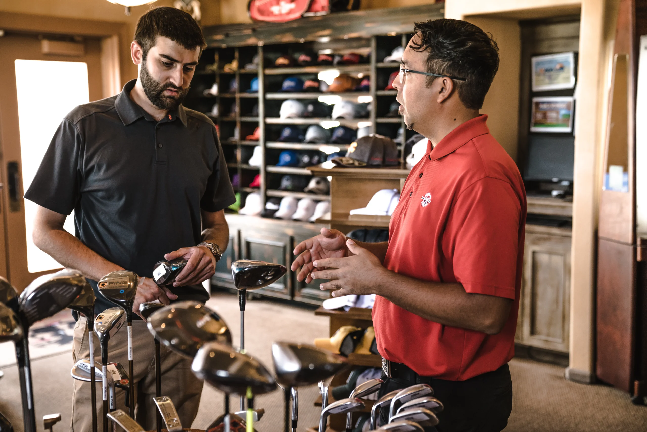 Two men discussing golf clubs in a pro shop, one holding a range finder.