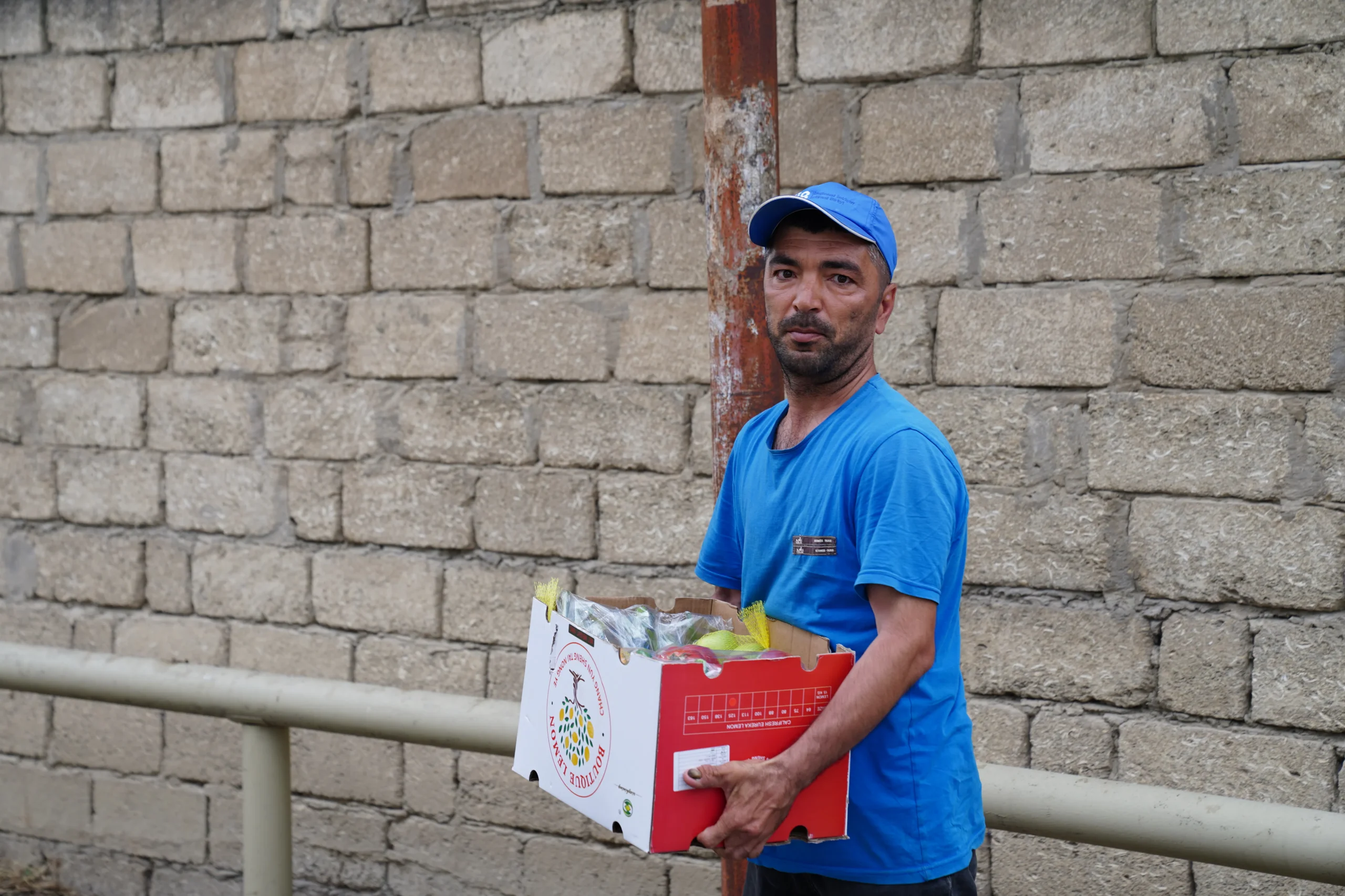 Man in blue carrying a box of produce in front of a brick wall.