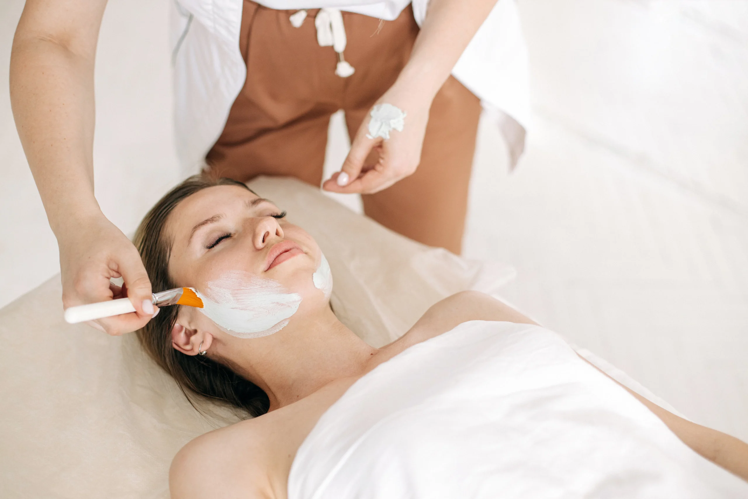 Woman receiving a facial with white mask applied by esthetician.
