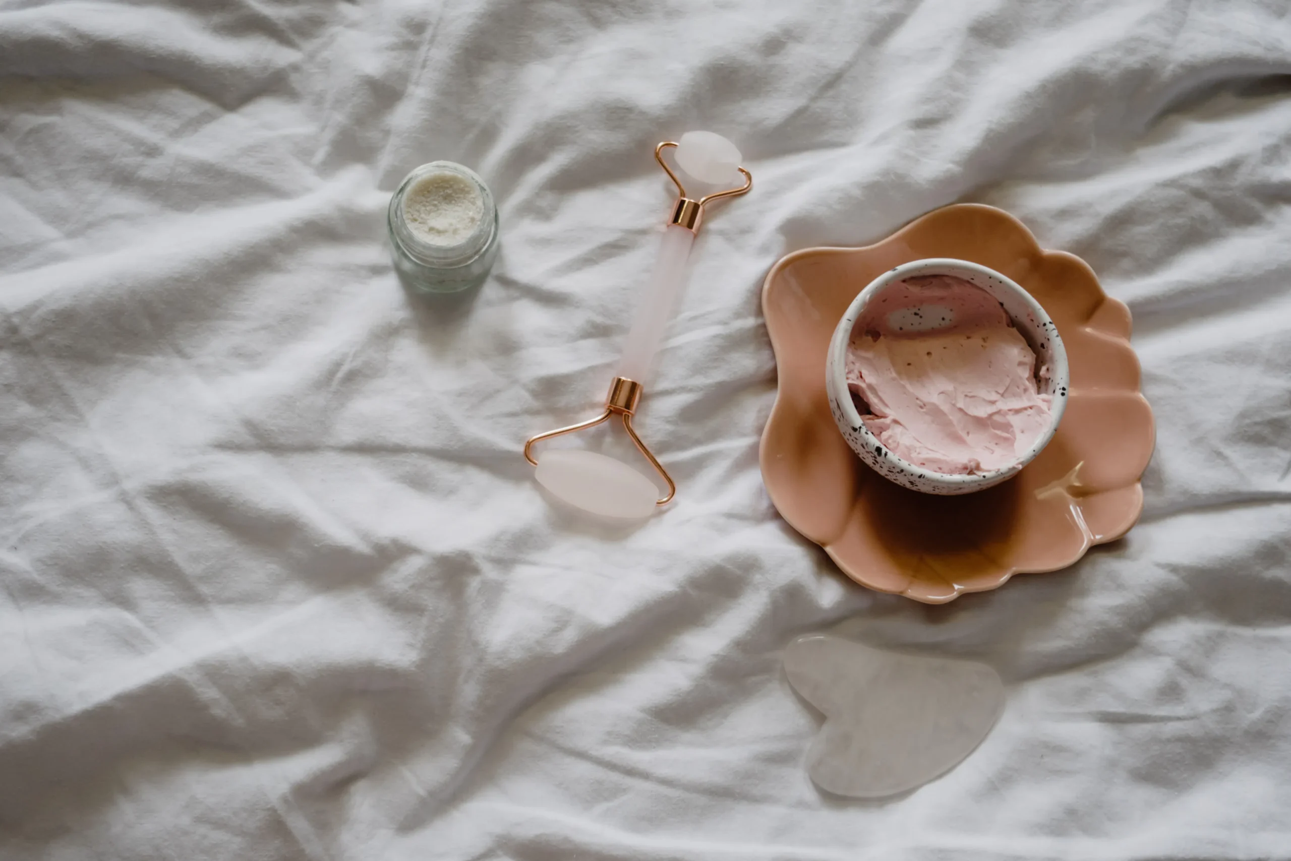 Skincare items on white wrinkled sheet: face roller, cream, gua sha, and bowl.