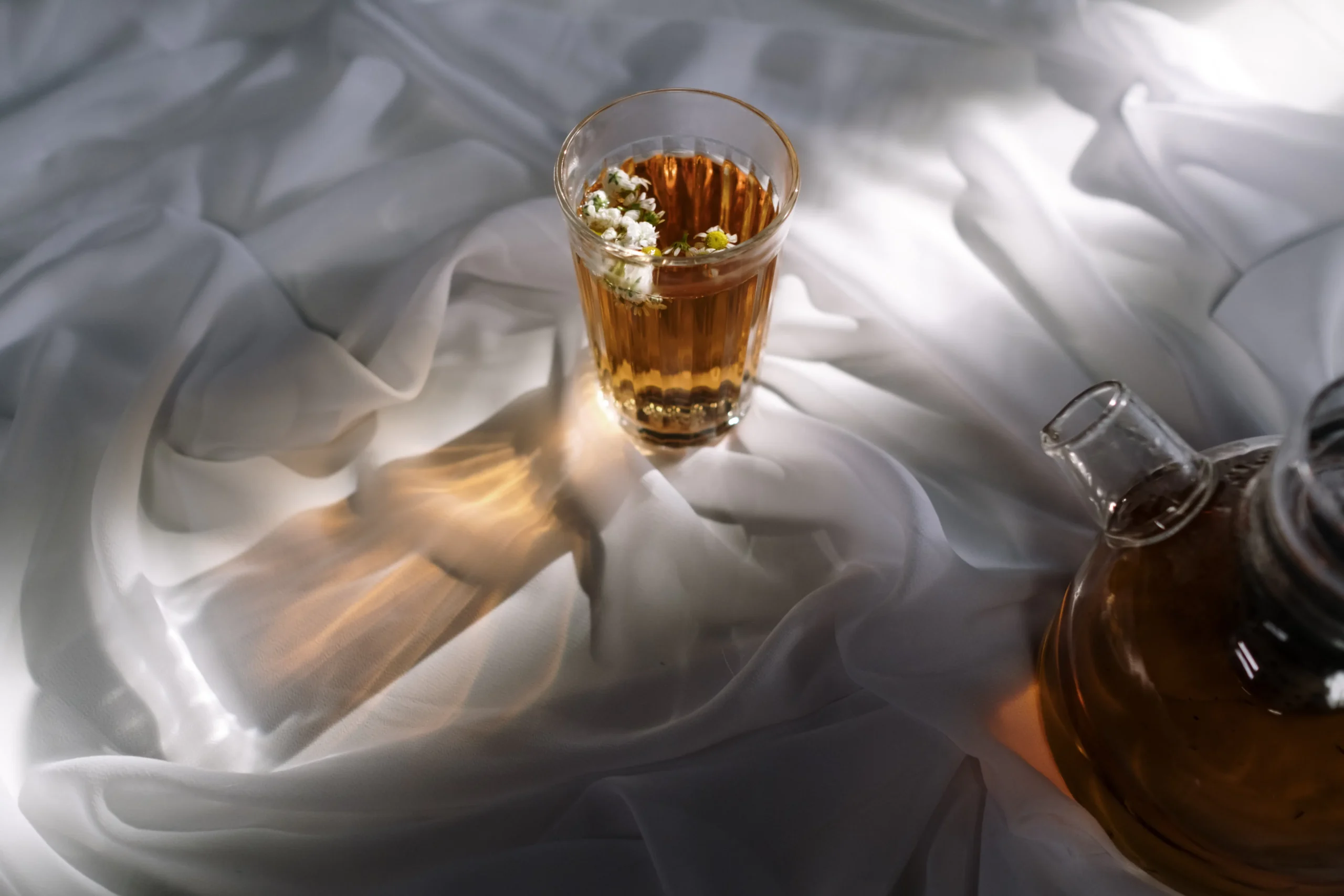 Glass of amber tea with white flowers and glass teapot on white draped fabric.