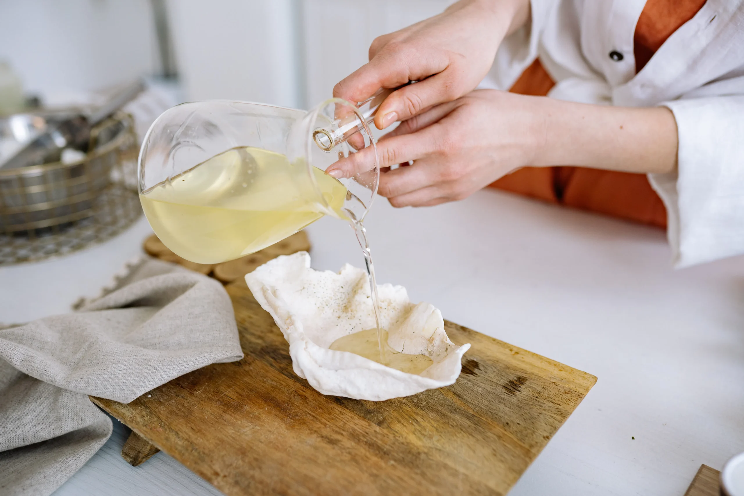 Person pouring yellow liquid from glass carafe into a white shell on a wooden board.