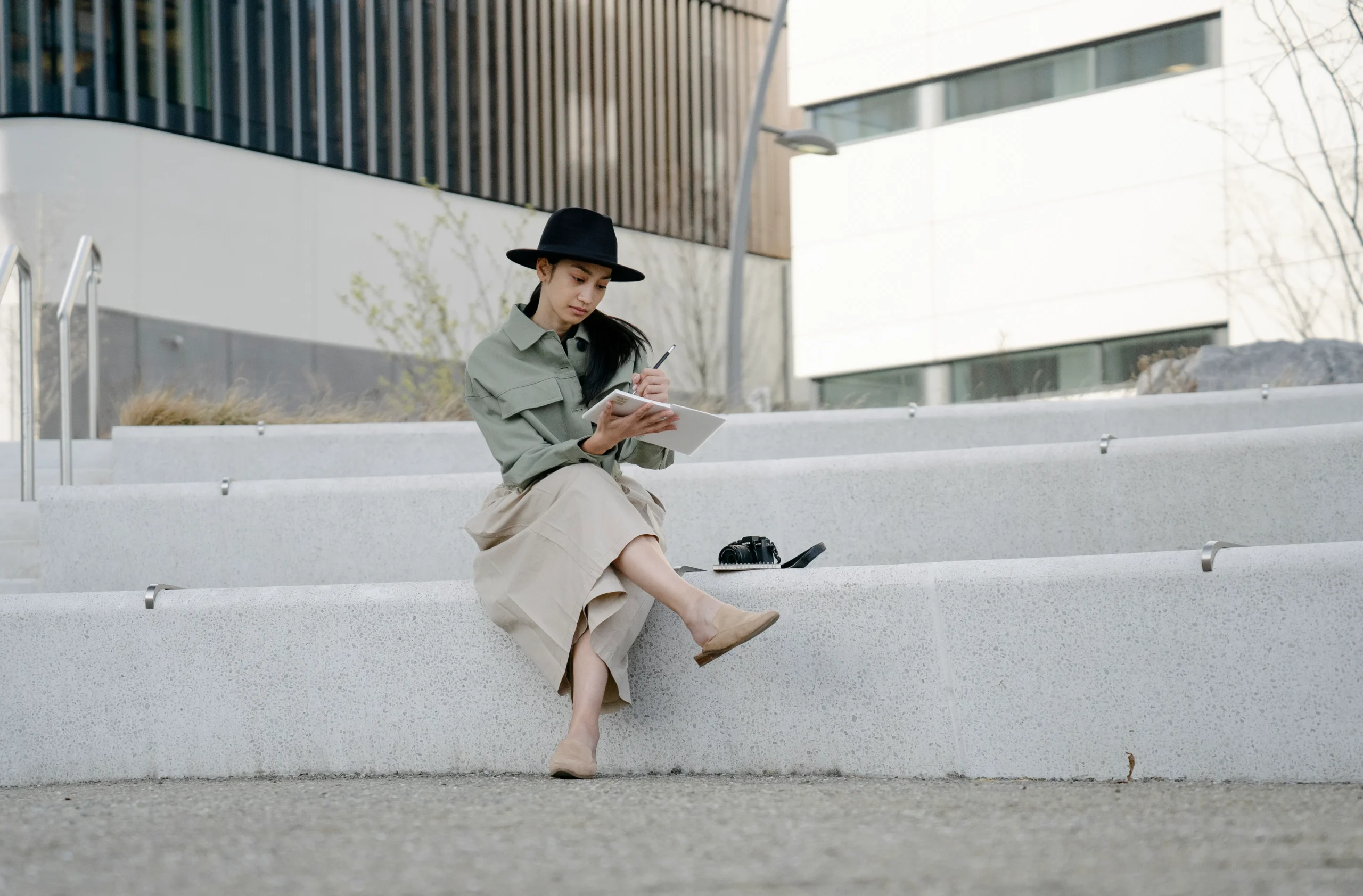 Woman in hat sitting on stairs, sketching in a notebook with a pen.