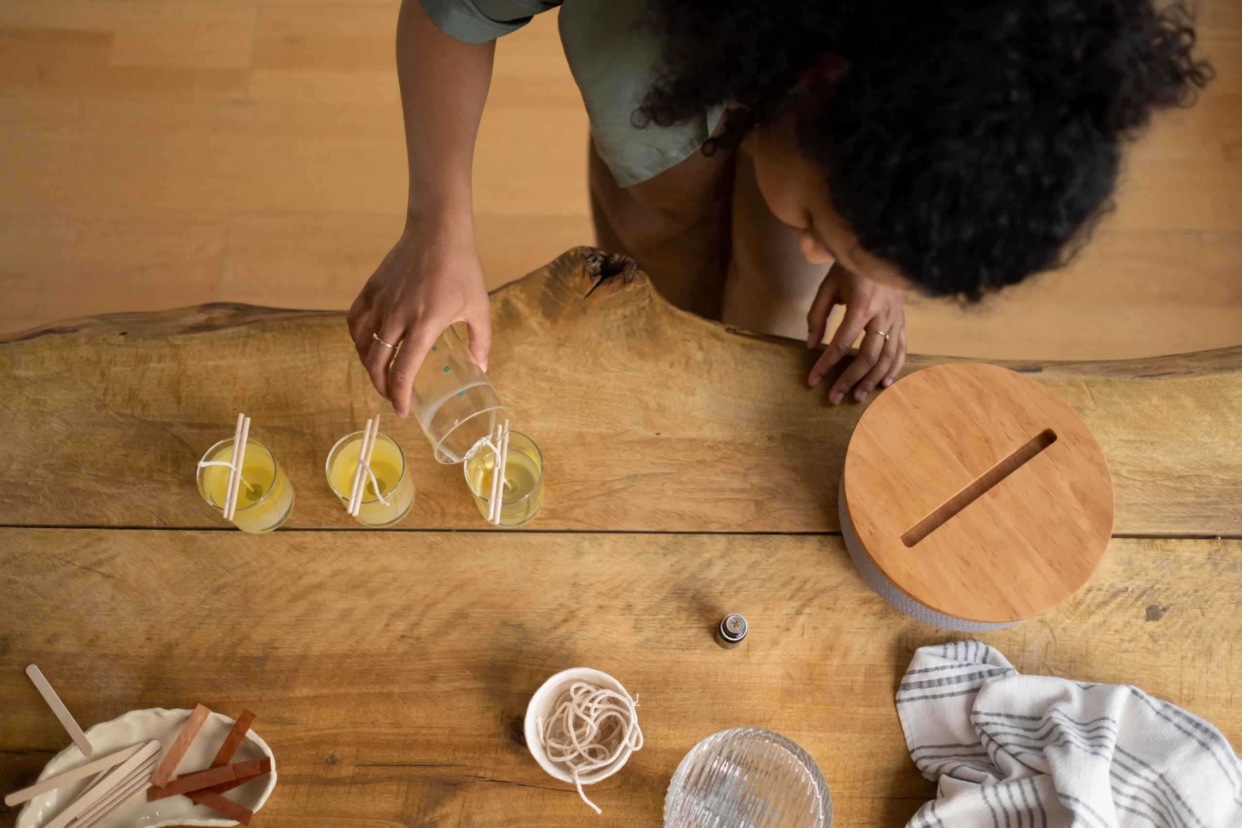 Overhead shot of a woman pouring wax into candle jars on a wooden table.