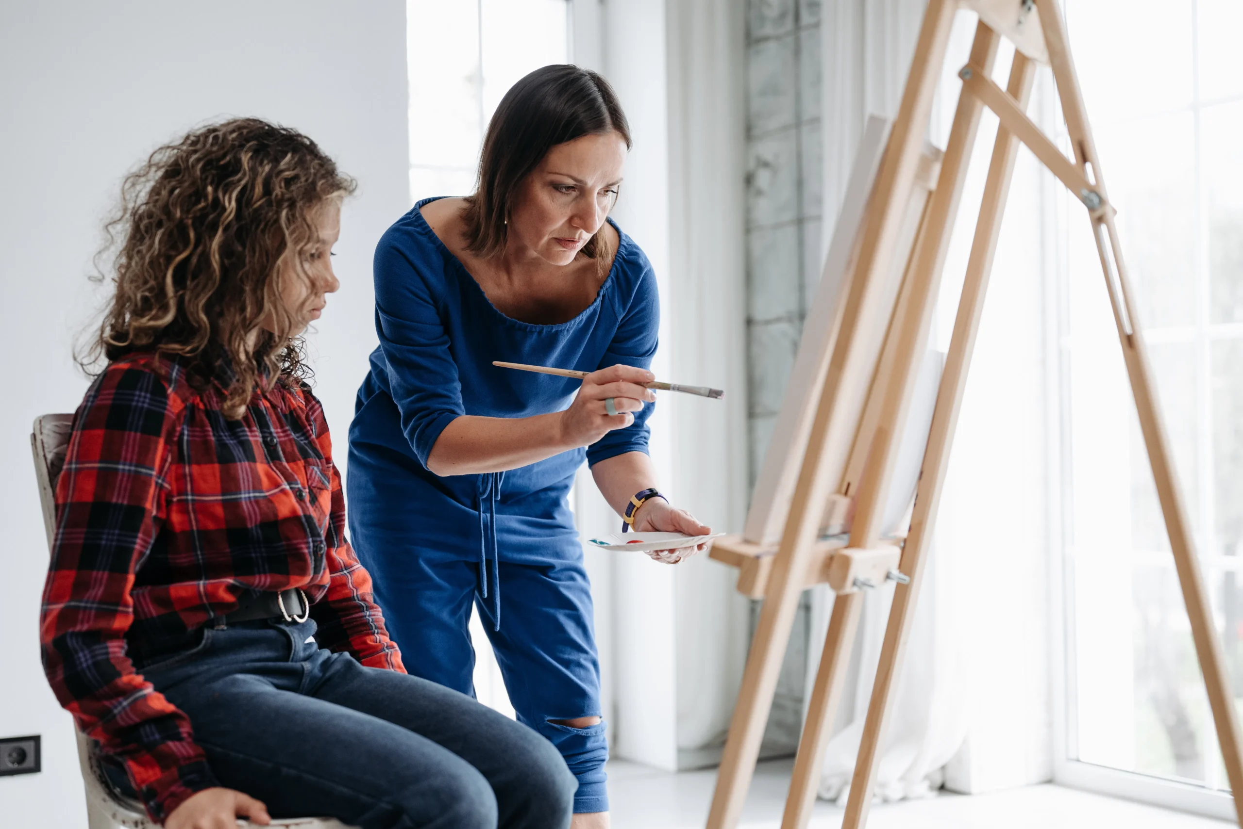 Woman guiding girl painting at easel, no connection to waxing context.
