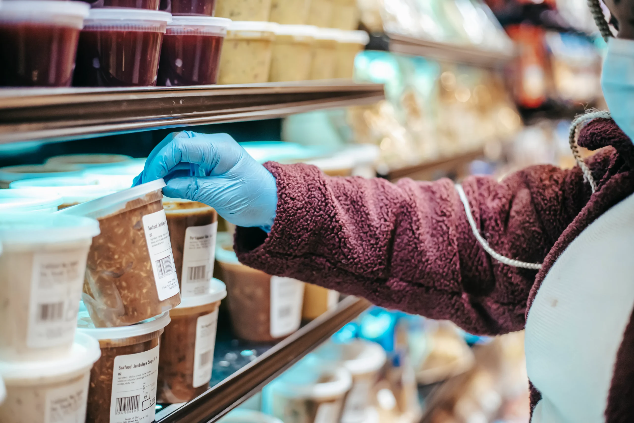 Gloved hand selecting packaged food from a grocery shelf, face mask visible.