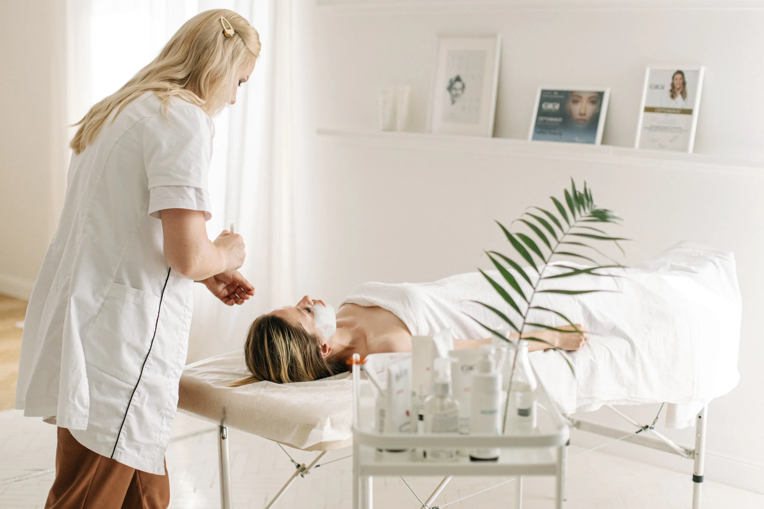 Blonde aesthetician in a white coat applies a facial treatment to a woman on a bed, representing popular skin rejuvenation options and carbon laser facial benefits in Amsterdam.