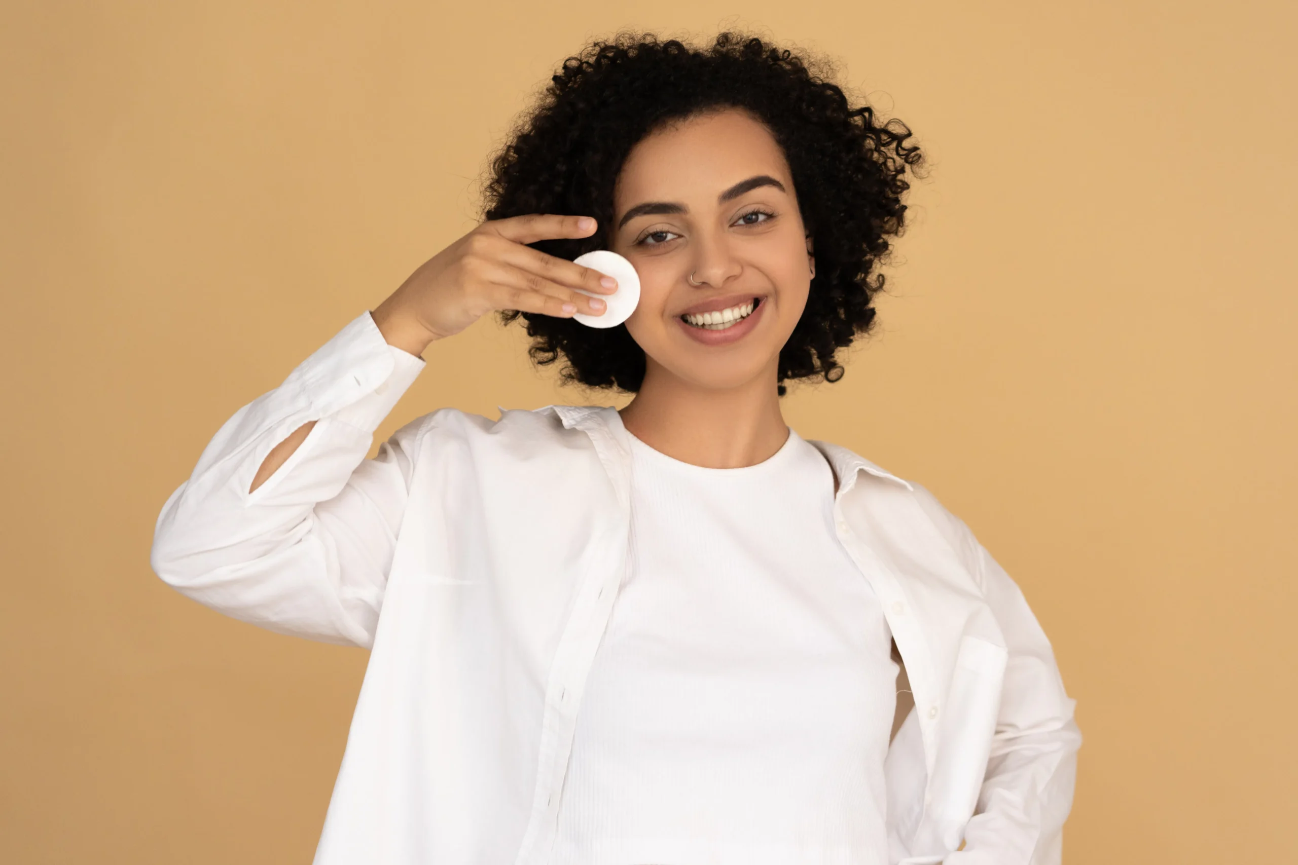Smiling woman with curly hair applying a cotton pad to her cheek, highlighting skincare treatments for a facial in Amsterdam.